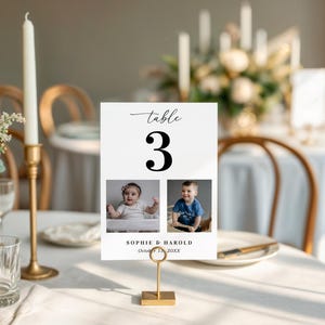 K&ouml;nnte beinhalten: Eine wei&szlig;e Tischnummerkarte mit der Zahl 3 in schwarzer Schrift. Die Karte zeigt zwei Fotos von Babys und die Namen "Sophie & Harold" mit dem Datum "15. Oktober 20XX". Die Karte wird von einem goldenen Metallst&auml;nder gehalten.