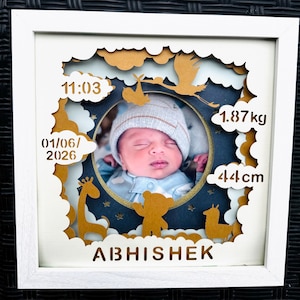 May include: A framed baby photo with a white border. The photo is surrounded by a layered design with gold-colored cutouts of clouds, animals, and the baby's name, Abhishek. The frame includes the baby's birth date, time, weight (1.87 kg), and length (44 cm).