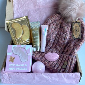 May include: A pink gift box filled with various items, including a pink knit hat with a fluffy pom-pom, a bath bomb, a pink eye mask, a mini puzzle, and two tubes of skincare products. The box is lined with pink tissue paper and has a brown cardboard exterior.