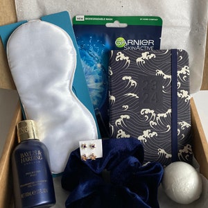 May include: A gift box filled with self-care items. Includes a white satin eye mask, a Garnier face mask, a navy blue journal with a wave pattern, a bottle of Baylis & Harding shower gel, a navy scrunchie, star earrings, and a bath bomb.