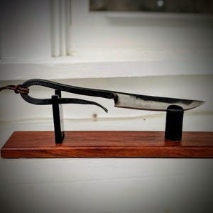 May include: A hand-forged, black metal knife with a curved blade, displayed on a wooden stand. The knife features a loop handle and a leather cord. The stand is made of dark wood and black metal supports.