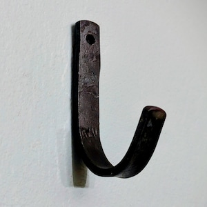 May include: A dark brown, hand-forged metal hook with a curved design. The hook has a rectangular top with a hole for mounting and a curved lower section. The metal has a textured surface and a rustic appearance, suitable for hanging items.