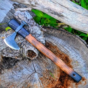 May include: A hand-forged tomahawk featuring a dark metal axe head with a curved blade and a hammer poll. The handle is crafted from polished brown wood, secured with a black leather wrap featuring visible stitching near the axe head.
