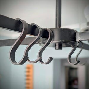 Puede incluir: Un conjunto de ganchos en S de metal negro colgados de un marco rectangular de metal negro. Los ganchos presentan una apariencia curva forjada a mano con un acabado mate. El marco está unido a un soporte central circular de metal negro.