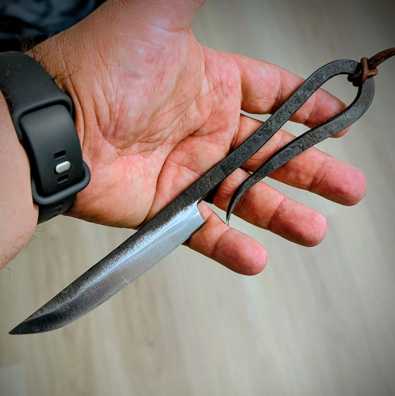 May include: Hand-forged knife with a dark, textured metal handle and a sharp, silver blade. The handle features a looped design with a leather cord for hanging. The blade is approximately 6 inches long.