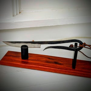 May include: A hand-forged knife with a dark, textured blade and handle, displayed on a red-toned wooden base. The knife is supported by a black cylindrical post and a curved metal stand. A leather cord is attached to the handle.