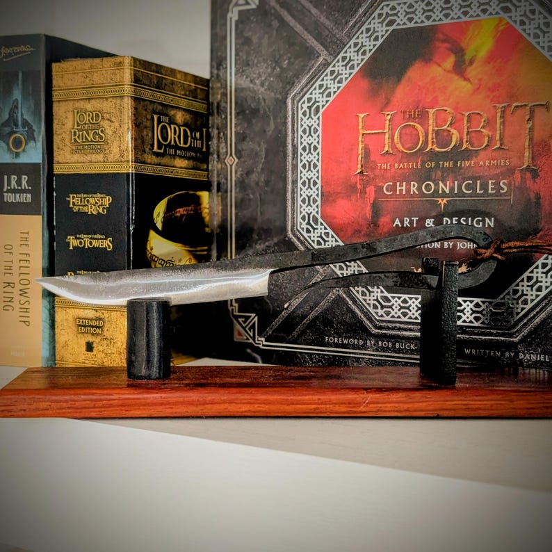 May include: A handcrafted metal knife with a silver blade and a dark, textured handle, displayed on a wooden shelf. The knife rests on a black stand, with several books in the background, including "The Hobbit" and "The Lord of the Rings" series.