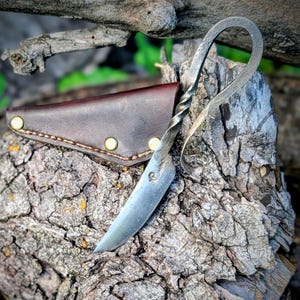 May include: A hand-forged steel knife featuring a twisted metal handle that loops into a circular pommel. The blade has a small stamped mark near the bolster. It is accompanied by a dark brown leather sheath secured with three brass rivets.