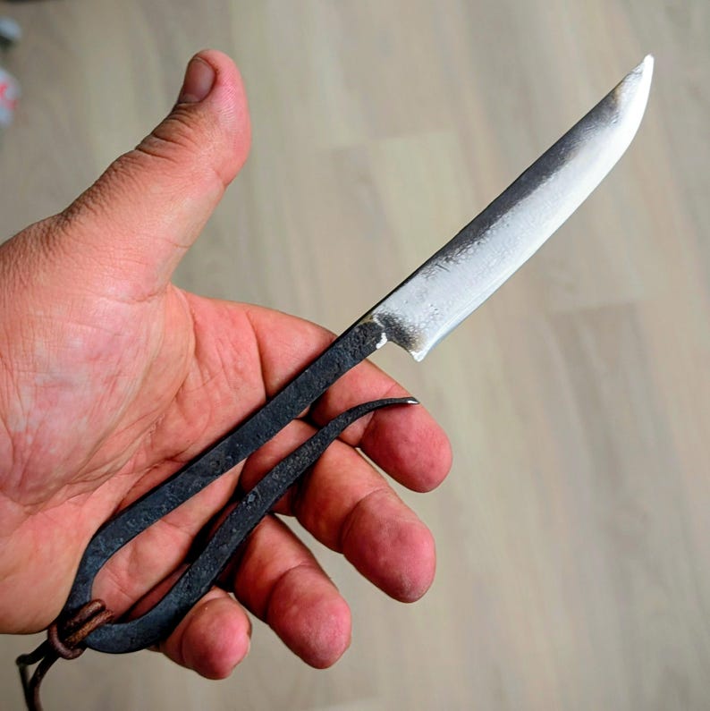 May include: Hand-forged knife with a curved handle and a sharp, silver blade. The handle is dark, possibly blackened steel, and features a loop at the end. A leather cord is attached to the loop. The knife is held in a hand.