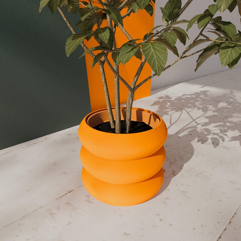 May include: A vibrant orange, three-tiered planter with a plant featuring green leaves and brown stems. The planter is set against a white surface with a dark green and orange background. The design is modern and minimalist, suitable for indoor or outdoor use.