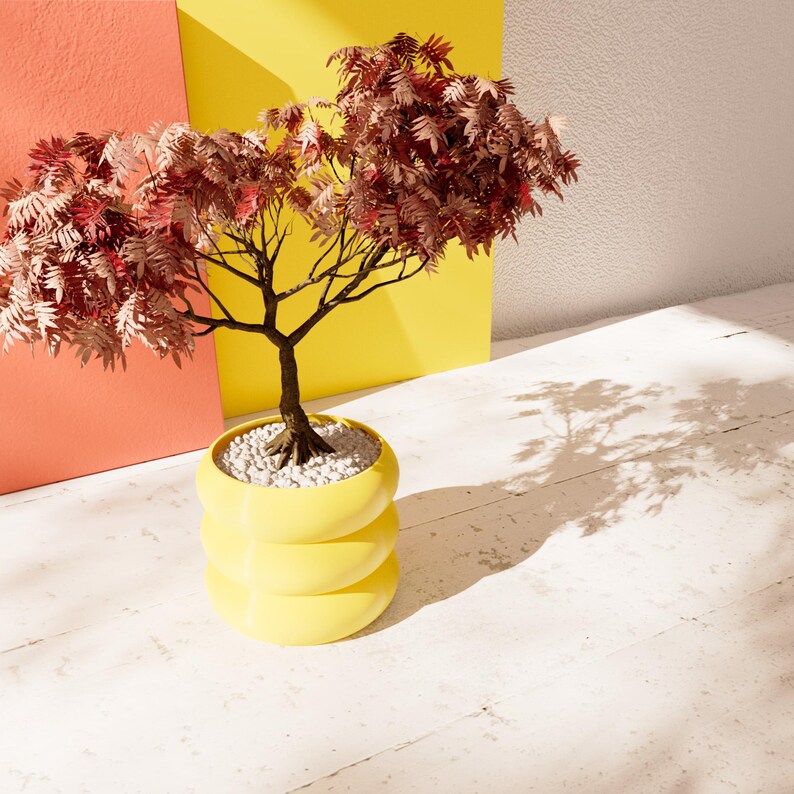 May include: A small tree with red-brown leaves in a yellow planter with three tiers. The tree is sitting on a white surface with a shadow cast from the tree.