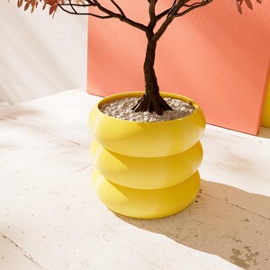 May include: A small tree with brown branches and red leaves is planted in a yellow, three-tiered planter. The planter is made of smooth, rounded, stacked sections.