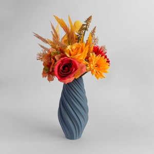 May include: A blue ceramic vase with a swirling design holds a bouquet of orange, yellow, and red flowers. The flowers include roses, gerbera daisies, and other fall-colored blooms.