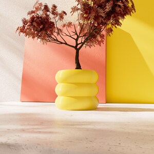 May include: A small tree with brown leaves in a yellow ceramic planter with three stacked rings. The planter is sitting on a white surface in front of a pink and yellow wall.