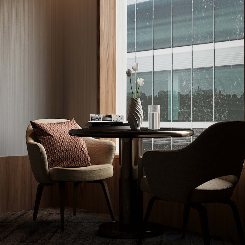 May include: A modern living room with two chairs and a table. The table has a vase with white flowers and a glass of water. The chairs are upholstered in a beige fabric. The room has a large window with a view of a city.