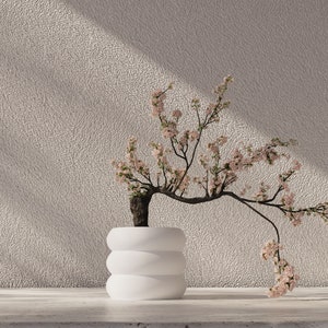 May include: A white ceramic planter with a rounded, layered design holds a delicate branch of a flowering tree with pink blossoms. The tree is set against a textured, light beige wall.