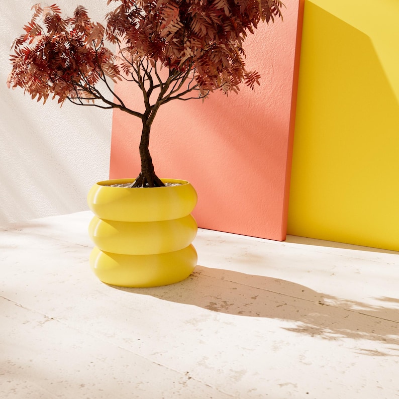 May include: A yellow ceramic planter with three tiers, featuring a small tree with brown leaves against a red and yellow background.