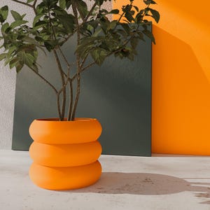 May include: An orange, three-tiered planter holds a leafy green plant. The planter is set against a backdrop of a dark green panel and an orange wall. The planter's design is modern and minimalist, suitable for indoor or outdoor use.
