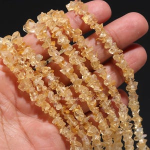 Op de afbeelding: Close-up van strengen ruwe, goudgele citrien edelsteen chips. De chips zijn onregelmatig van vorm en grootte, geregen om een ketting of armband te vormen. De edelstenen worden in een hand gehouden tegen een donkere achtergrond.