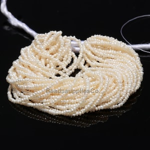 Op de afbeelding: Een streng witte zoetwaterparels, ongeveer 2 mm groot, met een licht onregelmatige vorm. De parels zijn geregen op een wit draad en zijn klaar voor het maken van sieraden.