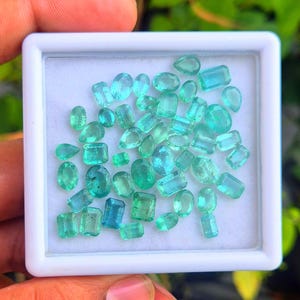 May include: A selection of emerald gemstones in a range of shapes, including oval, rectangular, and pear-cut. The gems are presented in a white square tray, displaying their vibrant green and teal colours. The image is well-lit, emphasising the clarity and brilliance of the stones.