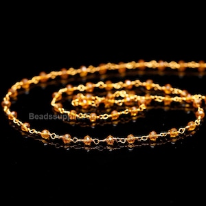 Könnte beinhalten: Eine goldene Kette mit kleinen, facettierten bernsteinfarbenen Perlen. Die Kette liegt auf einer schwarzen Oberfläche.