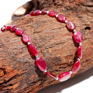 May include: A strand of ruby red gemstone beads with silver spacers, displayed on a piece of weathered wood.