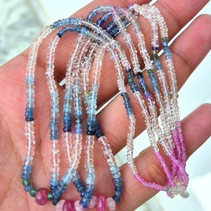 Könnte beinhalten: Eine Sammlung zarter Edelstein-Halsketten mit einer Vielzahl von Farben. Die Halsketten zeigen klare, blaue und rosa Perlen, wobei der Fokus auf den filigranen Details des Schmucks liegt. Die Perlen sind aufgefädelt und erzeugen einen mehrschichtigen Effekt.