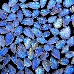 May include: A collection of carved gemstone leaves in shades of blue and white, scattered against a black background. The leaves have detailed veining and a translucent quality, creating a shimmering effect. The gemstones are likely used for jewelry making.