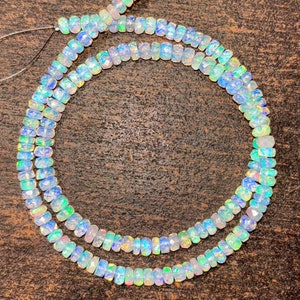 Puede incluir: Una hebra de cuentas de ópalo iridiscente en un esquema de colores azul claro, blanco y verde. Las cuentas son facetadas y tienen una forma ligeramente rectangular. La hebra está enhebrada en un hilo transparente y está lista para ser utilizada para la elaboración de joyas.