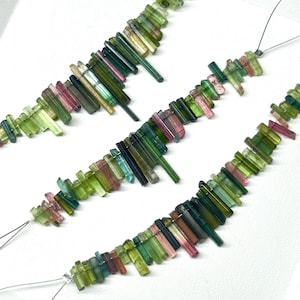 Pode incluir: Três fios de contas de turmalina verde, rosa e preta em pedras preciosas enfiadas em fio de prata. As contas são de tamanhos e formas diferentes e estão dispostas em um padrão em cascata.