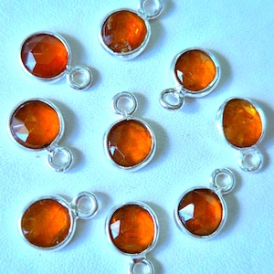 May include: A collection of eight silver-toned pendants with faceted, orange-colored gemstones. Each pendant has a circular or oval gemstone set in a silver bezel, with a small loop at the top for attaching to a chain or other jewelry component.
