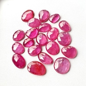 May include: A collection of faceted, oval-shaped rubies in varying sizes. The gemstones display a vibrant pinkish-red hue and are arranged on a white surface. The cut of the stones creates multiple reflective surfaces.