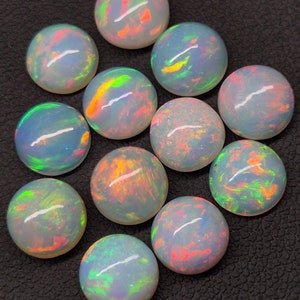 Może przedstawiać: Kolekcja dwunastu okrągłych, białych kaboszonów opalowych z tęczowymi, iryzującymi kolorami. Opale są ułożone w okrągły wzór na czarnym tle.