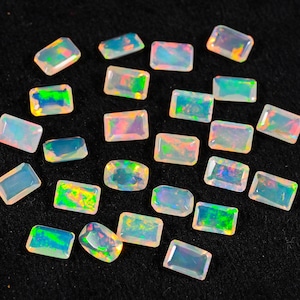 Puede incluir: Una colección de piedras preciosas de ópalo rectangulares con destellos iridiscentes de color verde, rosa y azul. Los ópalos están dispersos sobre una superficie negra, mostrando su apariencia pulida y translúcida. Estas piedras son ideales para la fabricación de joyas.