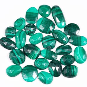 May include: A selection of polished malachite gemstones in diverse shapes, such as oval, round, and faceted. The stones exhibit a vivid green hue with characteristic dark green banding. The gemstones are displayed on a white background.