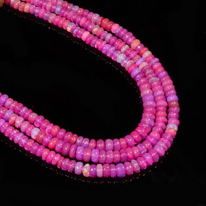 May include: A strand of pink opal beads, with a slight iridescent shimmer, is displayed on a black background. The beads are round and smooth, and they are arranged in a loose, flowing pattern.