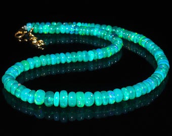 May include: A necklace composed of round, polished opal beads in shades of teal and turquoise. The beads are strung together, forming a continuous loop. The clasp is gold-colored. The necklace is displayed against a black background, highlighting its iridescent qualities.