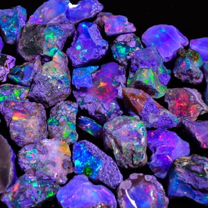 Puede incluir: Una colección de ópalos en bruto con colores vibrantes. Los ópalos muestran un espectro de colores, incluyendo azules, verdes y rojos, con un efecto brillante e iridiscente. Las piedras en bruto tienen una apariencia natural y texturizada, sobre un fondo oscuro.