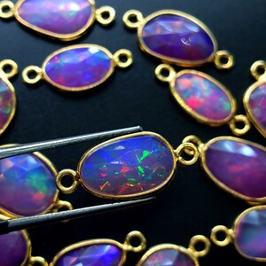 May include: A close-up of a collection of oval-shaped opal gemstones with gold-toned metal settings. The gemstones are iridescent with shades of purple, blue, and green. The gemstones are arranged in a loose pattern on a black surface.