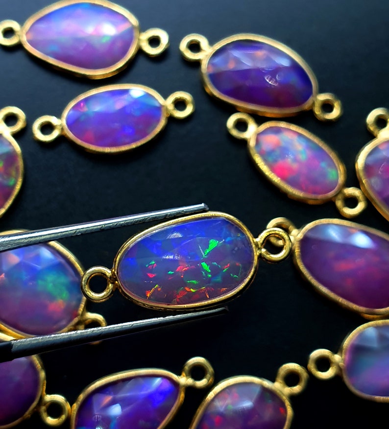 May include: A close-up of a collection of oval-shaped opal gemstones with gold-toned metal settings. The gemstones are iridescent with shades of purple, blue, and green. The gemstones are arranged in a loose pattern on a black surface.