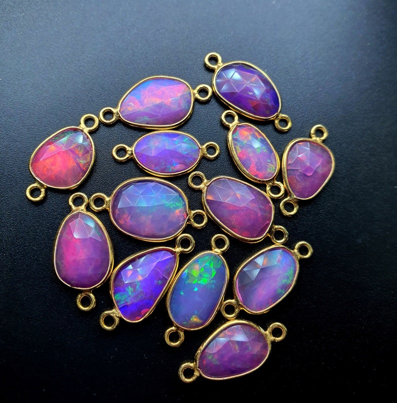 May include: A group of 15 iridescent opal gemstone connector charms with gold-tone metal loops. The opals are a mix of pink, purple, and blue.