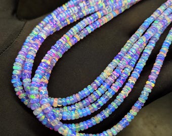 May include: Strands of iridescent opal beads in shades of blue, purple, and green. The beads are small and round, strung together to create multiple necklaces. The opals have a shimmering, colorful appearance, and are displayed against a dark background.