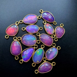 May include: A group of 15 oval-shaped gemstone pendants with gold-toned jump rings. The gemstones are iridescent and have a mix of pink, purple, and blue hues.