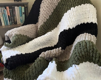 May include: A chunky knit throw blanket with stripes of white, black, green, and gray yarn. The blanket is draped over a white rocking chair.