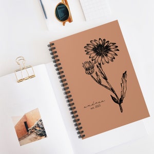 Puede incluir: Una libreta espiral marrón con un dibujo en línea negra de una margarita en la portada. El texto "andrea est. 2023" está impreso debajo de la flor.
