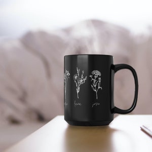 Puede incluir: Taza de caf&eacute; de cer&aacute;mica negra con un dise&ntilde;o floral blanco y los nombres "Jenna" y "Jesse" escritos debajo de las flores.