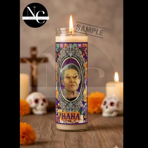 May include: A lit glass candle featuring a portrait of a woman in an ornate frame. The candle has a purple and floral design with the word "NANA" at the bottom. The word "SAMPLE" is printed across the top.