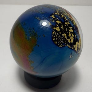 Puede incluir: Un globo terr&aacute;queo decorativo de cristal con un oc&eacute;ano azul y continentes con detalles de pan de oro. El globo est&aacute; montado sobre una base negra. Los continentes est&aacute;n detallados con pan de oro, y el oc&eacute;ano tiene patrones azules arremolinados.
