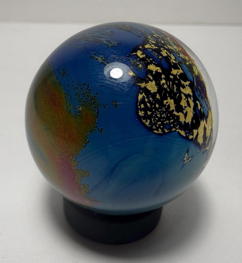 Puede incluir: Un globo terr&aacute;queo decorativo de cristal con un oc&eacute;ano azul y continentes con detalles de pan de oro. El globo est&aacute; montado sobre una base negra. Los continentes est&aacute;n detallados con pan de oro, y el oc&eacute;ano tiene patrones azules arremolinados.